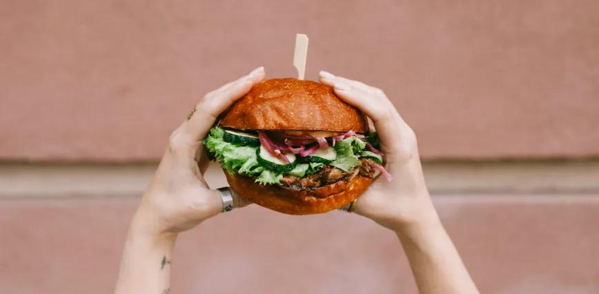 Two hands holding up a freshly made burger against a pink wall