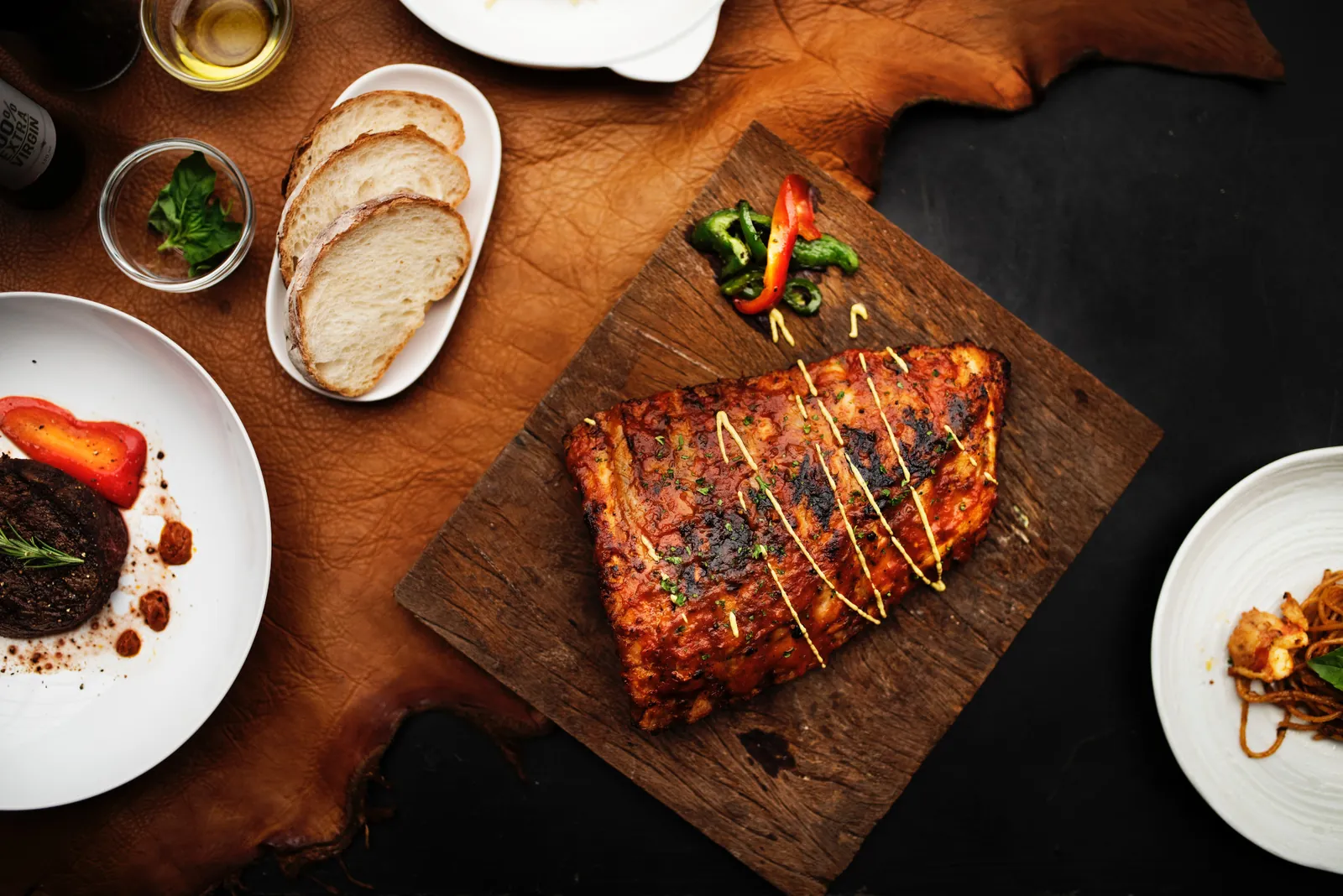 An overhead spread of glazed ribs on a carving board, alongside steak, sliced bread and olive oil