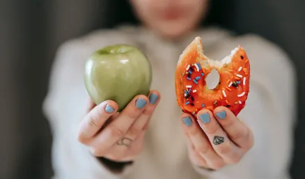 A person holding up a green apple in one hand and a frosted doughnut in the other